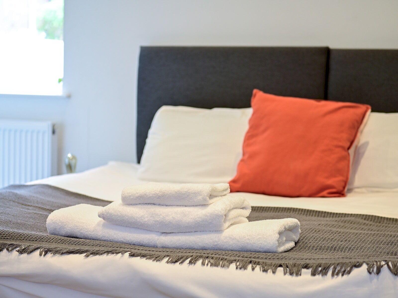 A neatly made bed with white and orange pillows, a grey headboard, and a grey and white striped blanket.
