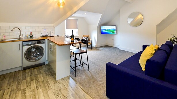 A modern apartment with a kitchen, dining area, and living room. It features a washing machine, a dining table with chairs, a sofa, and a flat-screen TV mounted on the wall. - Roath - Modern Flat Near City Centre W/Parking | Pass the Keys (Cardiff)