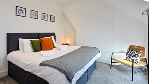A bedroom with a bed, pillows, a bedside table, a chair, and framed pictures on the wall. - Roath - Modern Flat Near City Centre W/Parking | Pass the Keys (Cardiff)