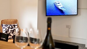 A room with a television displaying a boat on water, a wicker chair, and a table with glasses and a bottle. - Roath - Modern Flat Near City Centre W/Parking | Pass the Keys (Cardiff)