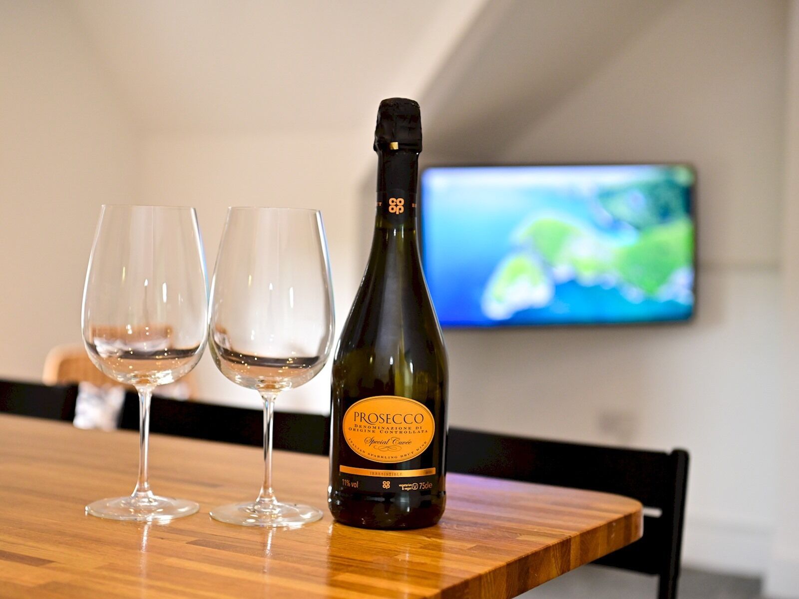 A bottle of Prosecco wine and two empty wine glasses on a wooden table with a TV in the background.