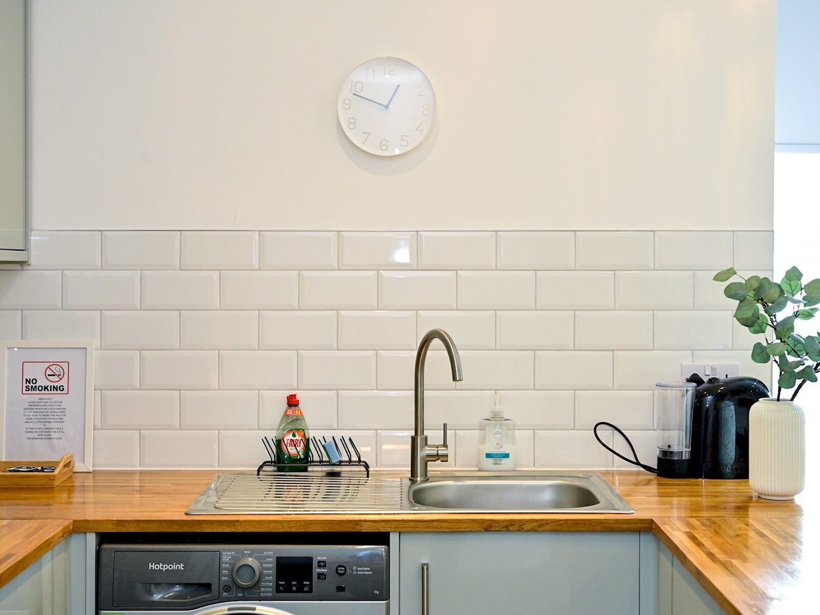 Une cuisine équipée d’un évier, d’une horloge, d’un égouttoir et d’un panneau indiquant qu’il est interdit de fumer.