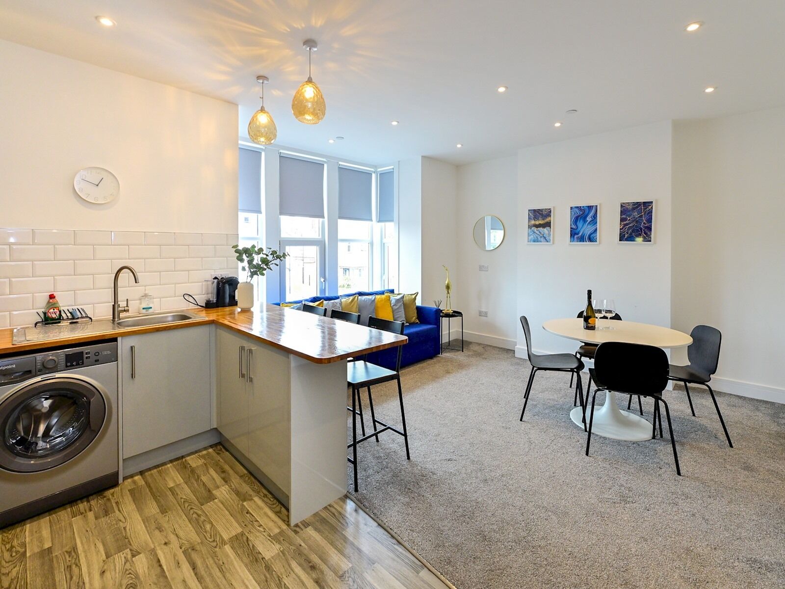A modern kitchen with a built-in washing machine, a dining area with a round table and chairs, and a blue sofa in a bright living space with large windows.