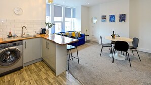 A modern kitchen with a built-in washing machine, a dining area with a round table and chairs, and a blue sofa in a bright living space with large windows.