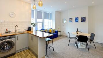 A modern kitchen with a built-in washing machine, a dining area with a round table and chairs, and a blue sofa in a bright living space with large windows.