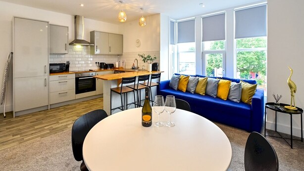 A modern kitchen with white cabinets, a wooden countertop, and a built-in oven. There is a dining area with a round table, chairs, and a bottle of wine. A blue sofa with colorful cushions is against a window with a view of greenery outside.