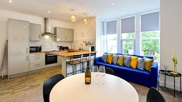 A modern kitchen with white cabinets, a wooden countertop, and a built-in oven. There is a dining area with a round table, chairs, and a bottle of wine. A blue sofa with colorful cushions is against a window with a view of greenery outside.