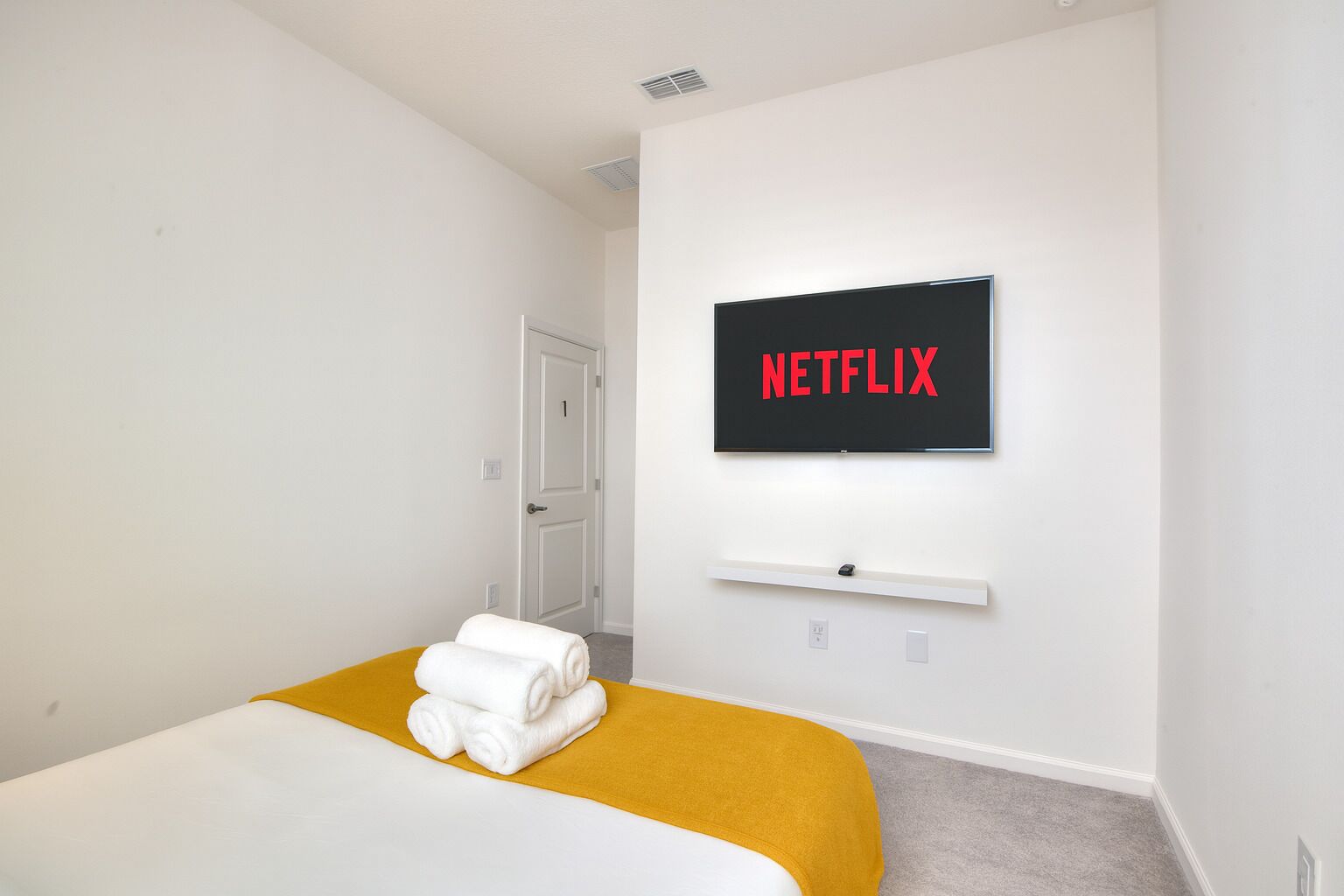 A hotel room with a bed, a TV mounted on the wall displaying 'NETFLIX', and a door leading to another room.