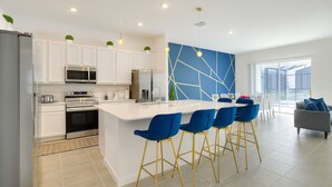 A modern kitchen with white cabinets, stainless steel appliances, and a blue accent wall. There are four blue bar stools with gold legs around a central island.