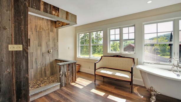 A bathroom with a wooden accent wall, a freestanding bench, a bathtub, and large windows with a view of greenery. - The Farmhouse at Pine Creek (Watson Township)