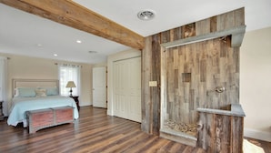 A bedroom with a wooden floor, a bed with a headboard, a bench, and a rustic wall with a built-in shelf. - The Farmhouse at Pine Creek (Watson Township)