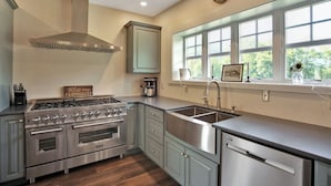 A kitchen with a gas stove, oven, sink, and dishwasher. There is a window with a view of greenery outside. - The Farmhouse at Pine Creek (Watson Township)
