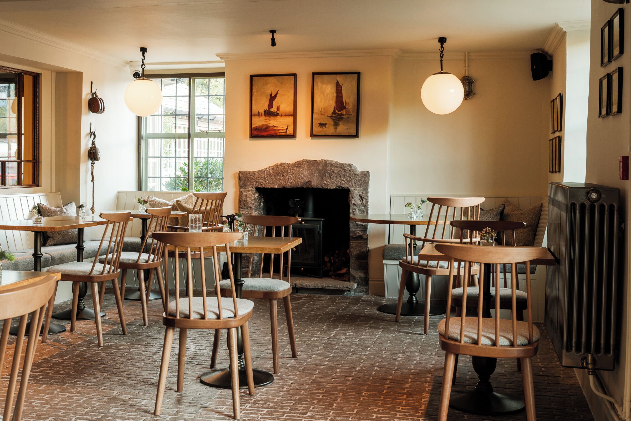 A cozy restaurant with brick flooring, wooden tables and chairs, a fireplace, and artwork on the walls.