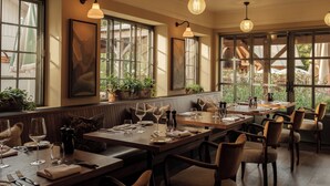 A restaurant with multiple tables, each set with wine glasses, plates, and cutlery. There are potted plants on the windowsill and a view of the outside through large windows. - The Royal George (Tintern)
