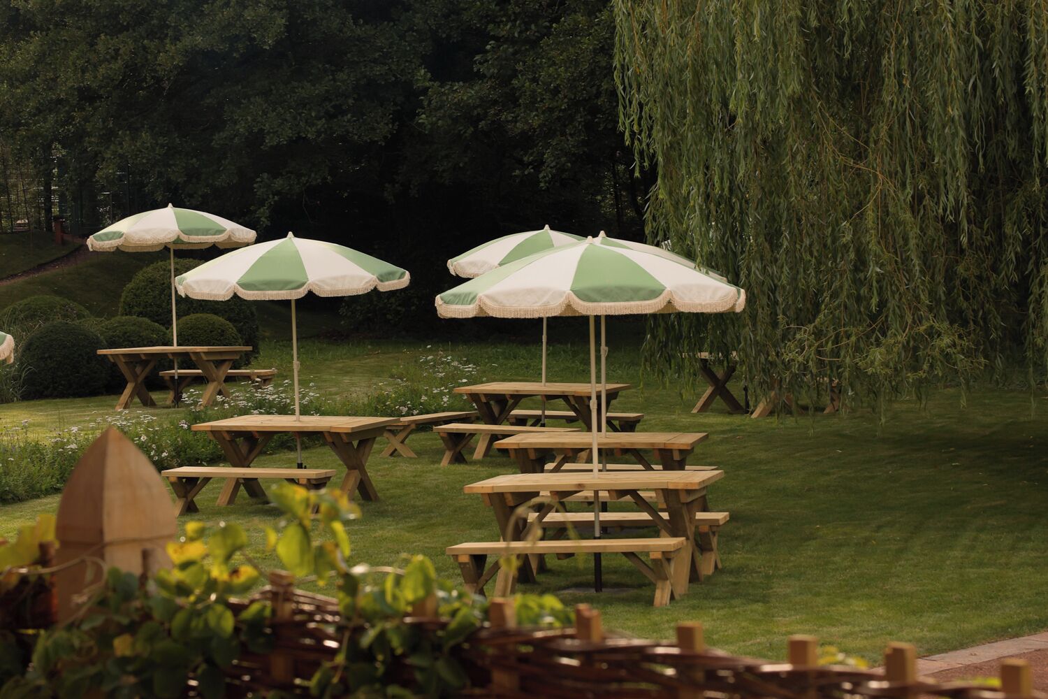 Outdoor wooden picnic tables with green and white striped umbrellas, arranged on a grassy lawn with trees in the background.