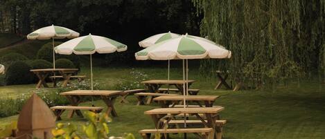 Outdoor wooden picnic tables with green and white striped umbrellas, arranged on a grassy lawn with trees in the background.