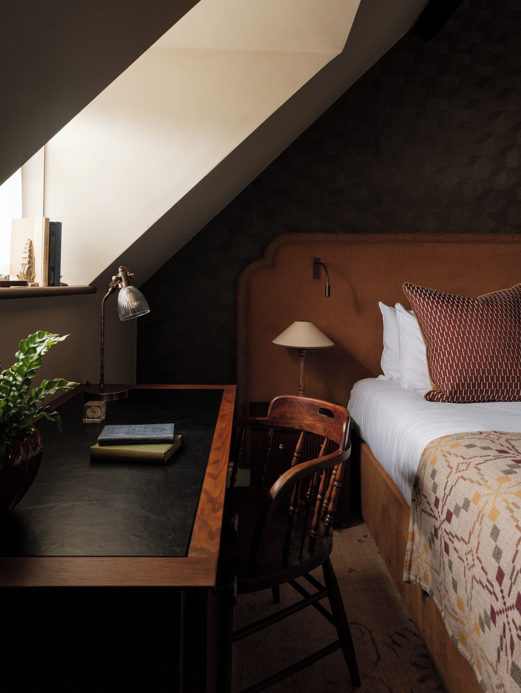 A cozy room with a wooden desk, a chair, a bed with patterned bedding, and a lamp.