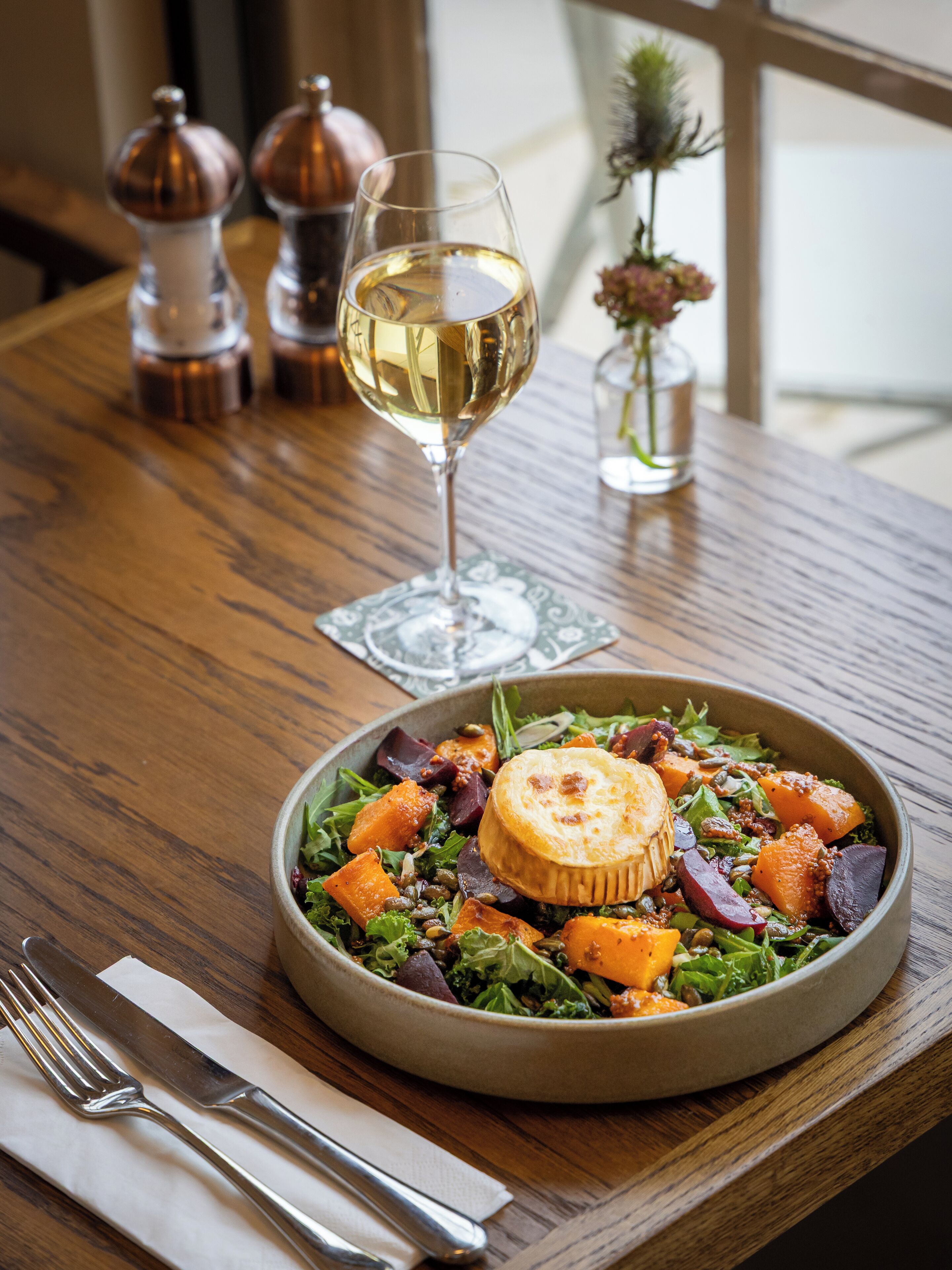 Une table en bois, une assiette de nourriture, un verre de vin blanc et un vase rempli de fleurs.