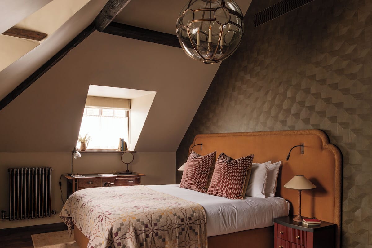 Ein Schlafzimmer mit schrägen Wänden, einem großen Bett mit gemusterten Textilien, einem Nachttisch mit Lampe und einem Fenster mit Blick ins Freie.