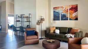 A spacious living room with a brown leather sofa, a round coffee table, and a green sofa with decorative pillows. There is a bookshelf with various items and a floor lamp.