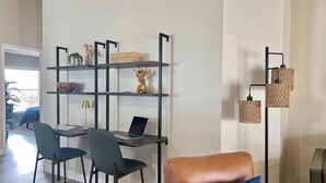 A modern living room with a brown leather sofa, a dining table with two chairs, a black metal shelving unit with decorative items, and a floor lamp.