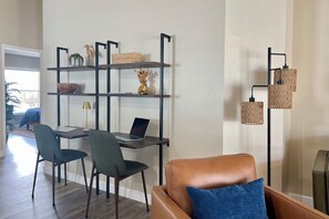 Una sala moderna con un sofá de cuero marrón, una mesa de comedor con dos sillas, un estante metálico negro con objetos decorativos y una lámpara de pie.