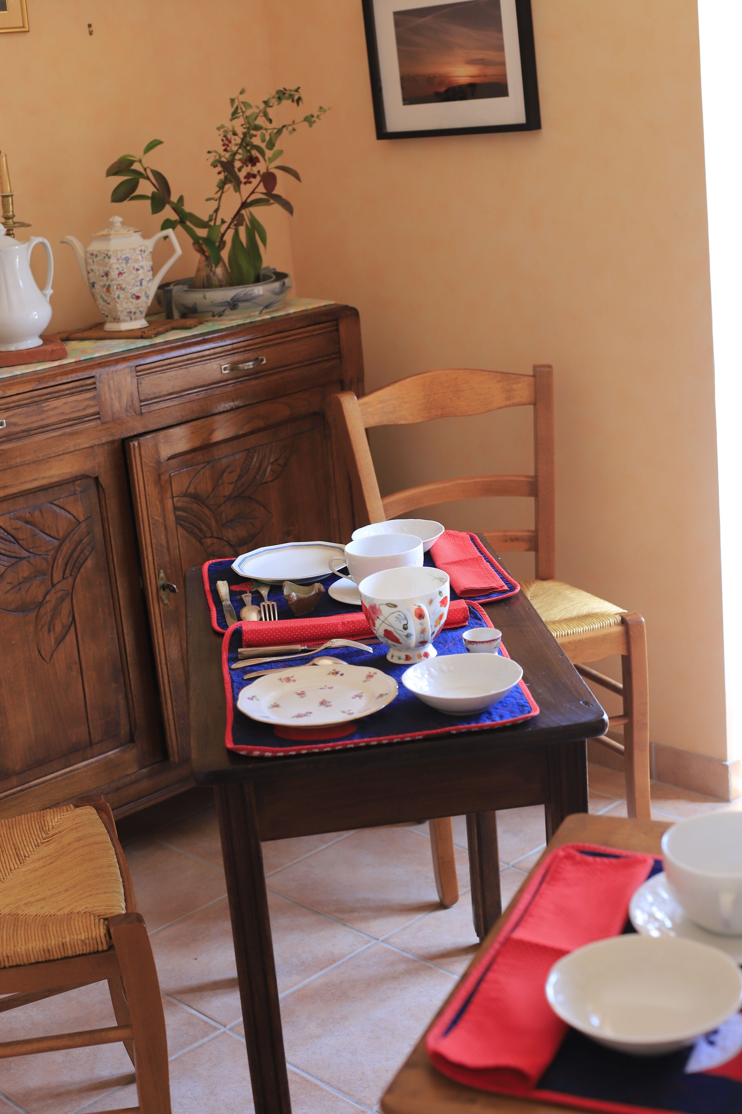 Taula de dinar preparada amb plats, cuplets i una tetera, amb una cadira i un armari de fusta al fons.