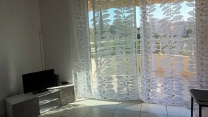 A room with a flat-screen TV on a stand, a sofa, and a chair. There is a window with a view of a building and trees. - House Swap Frejus (Frejus)