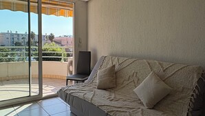 A modern living room with a grey sofa, a small table, and a chair. There is a sliding glass door leading to a balcony with a view of buildings and greenery. - House Swap Frejus (Frejus)