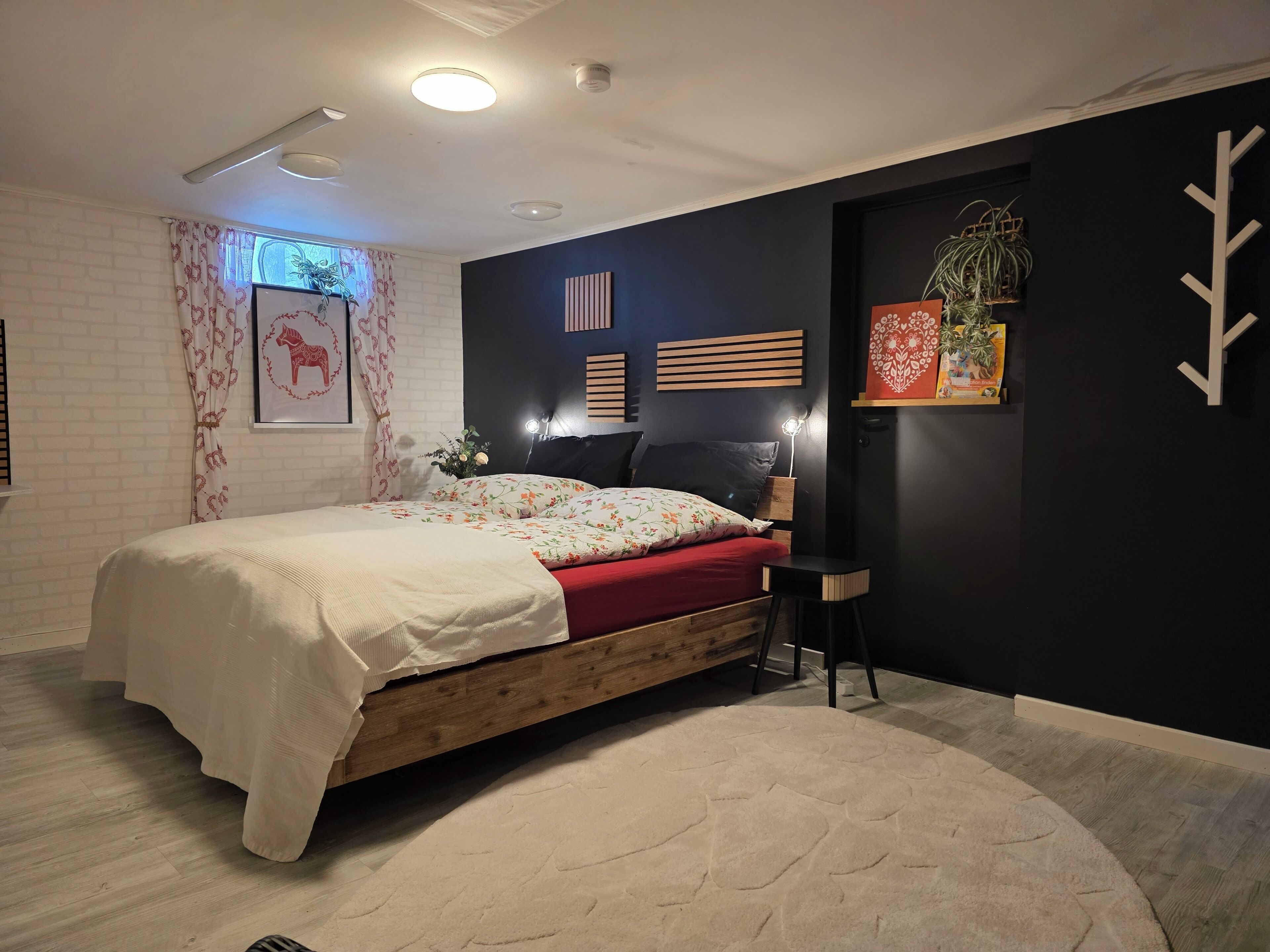 A bedroom with a bed, a nightstand, a chair, a wall-mounted shelf with plants, and a window with curtains.