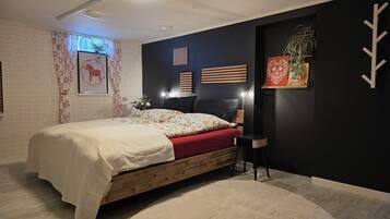 A bedroom with a bed, a nightstand, a chair, a wall-mounted shelf with plants, and a window with curtains.