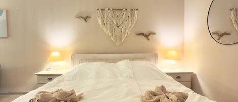Una cama bien hecha con toallas dispuestas en un diseño decorativo, un adorno de macramé en la pared y dos lámparas de pared con luces con forma de pájaro.