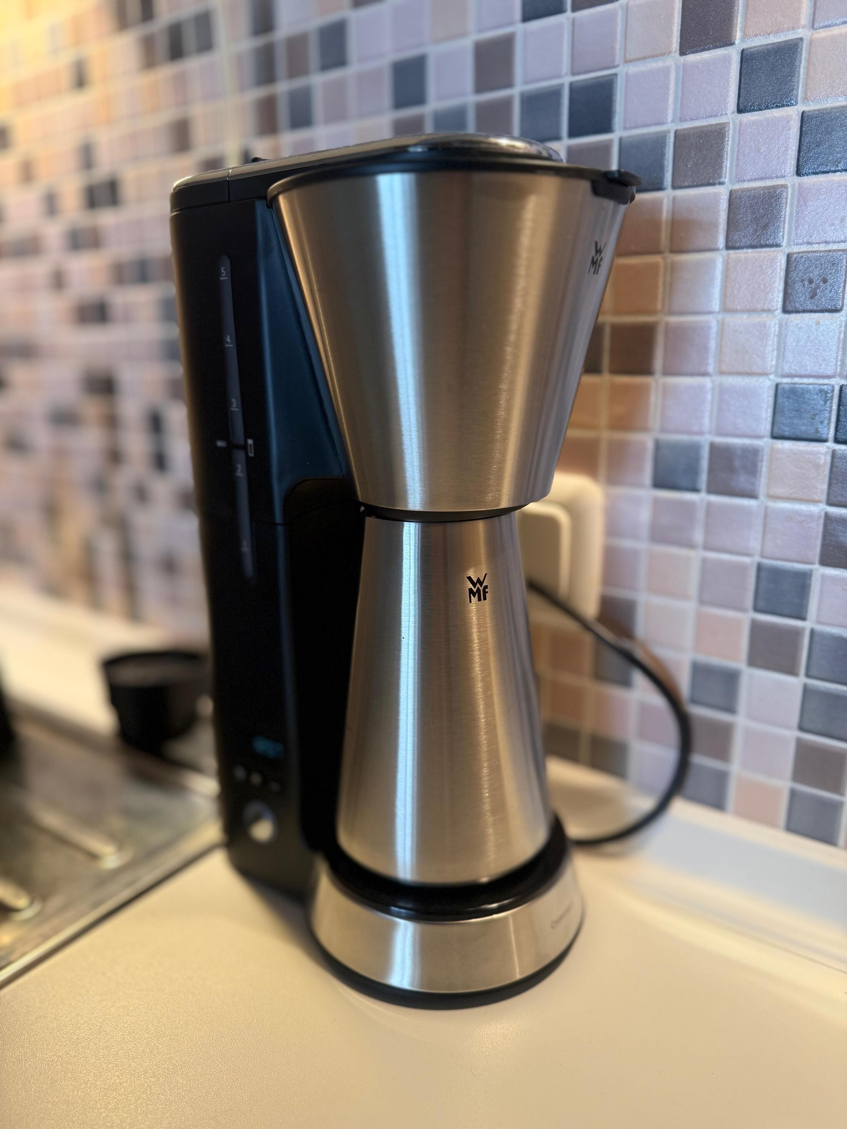 A coffee maker with a metallic finish, connected to a black device, against a tiled backsplash.