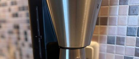 A coffee maker with a metallic finish, connected to a black device, against a tiled backsplash.
