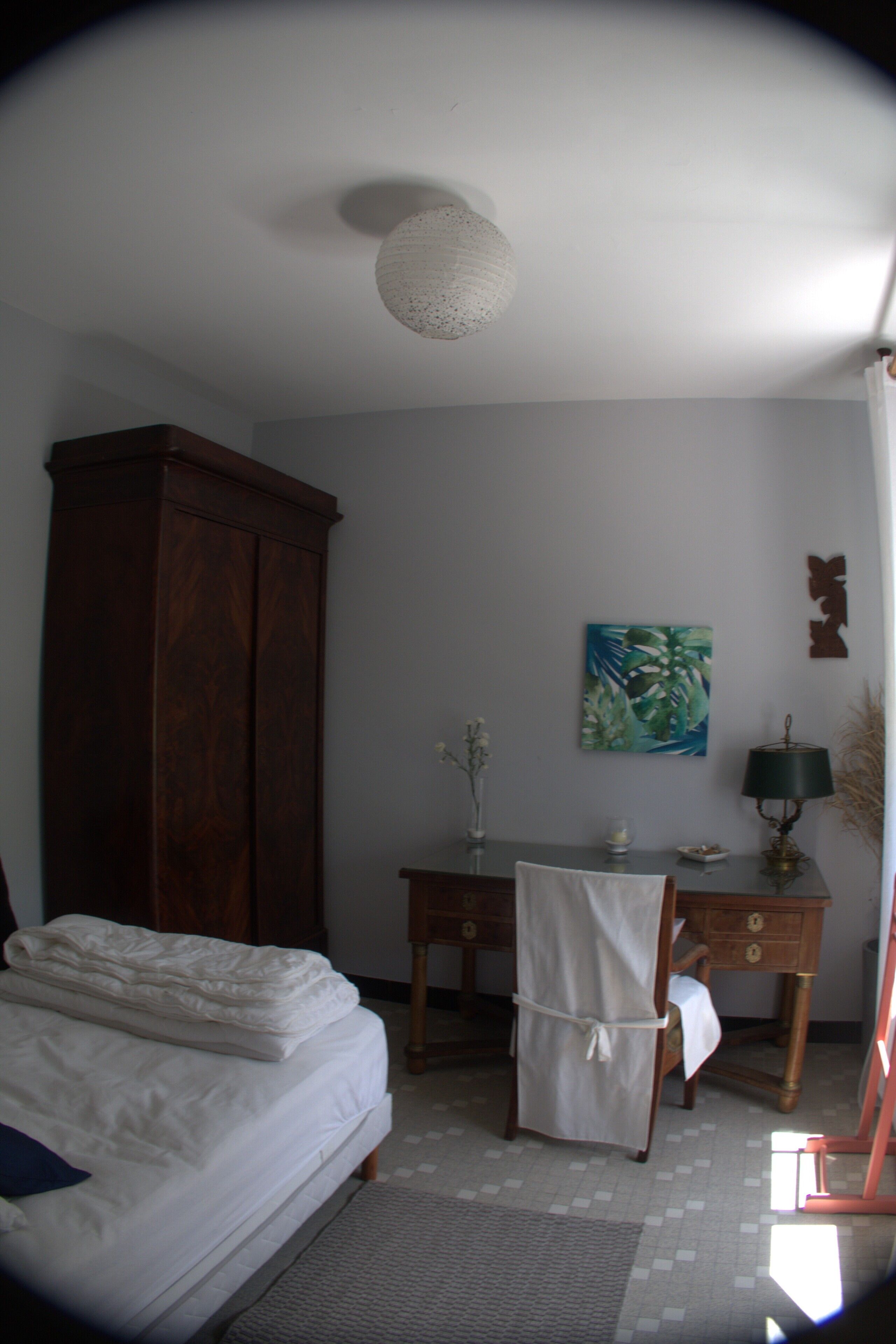 A bedroom with a bed, a wooden wardrobe, a desk with a lamp, and a painting on the wall.