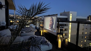 Une terrasse sur le toit équipée d’un grand écran diffusant un film, un coin salon confortable avec des coussins, et une vue sur le paysage urbain au crépuscule.