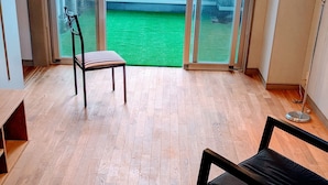 A room with wooden flooring, a black sofa, a striped chair, and a view through large glass doors to a green outdoor area.