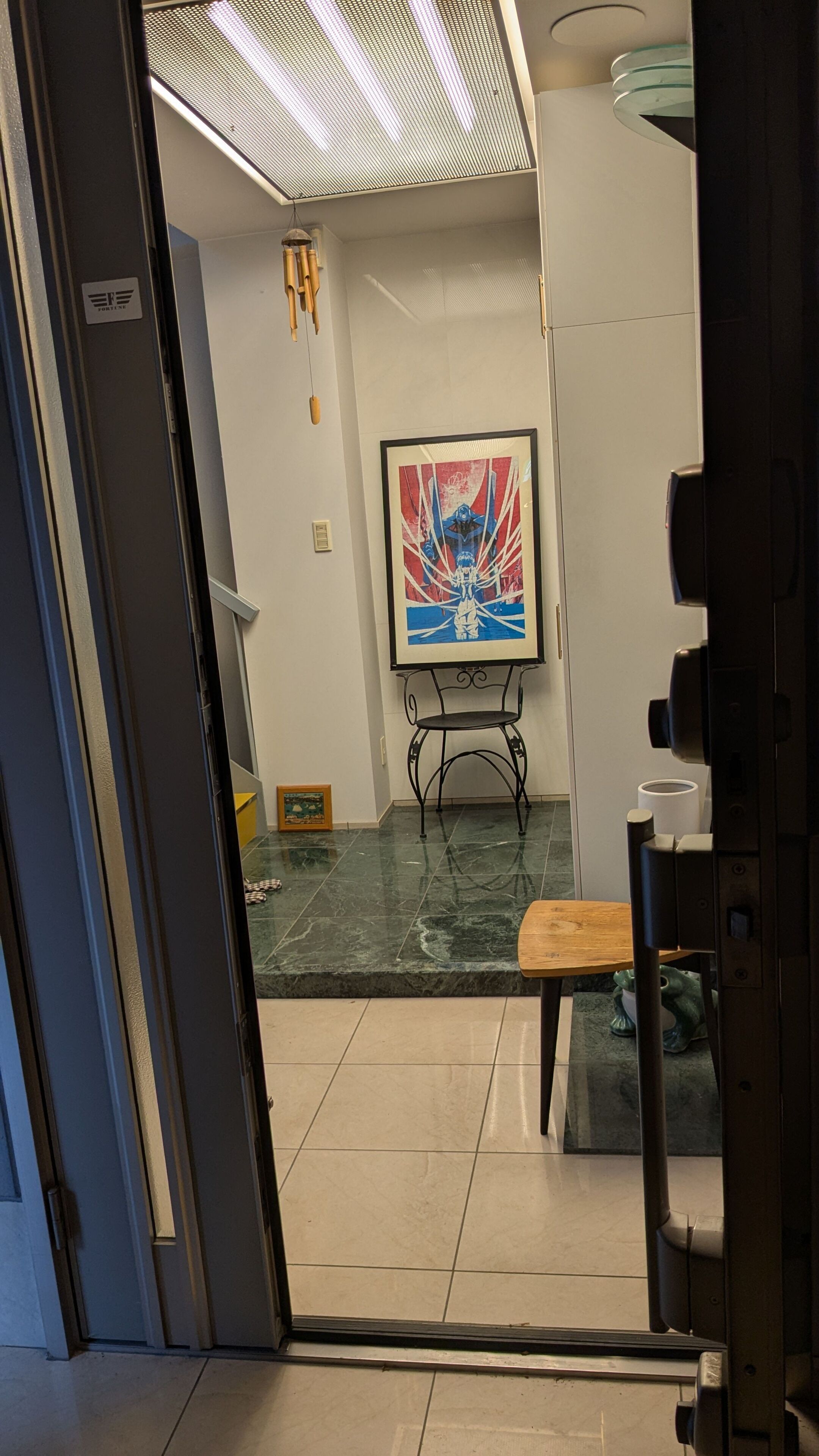 An interior view of a room with a tiled floor, a framed artwork on the wall, a small wooden table, and a chair.