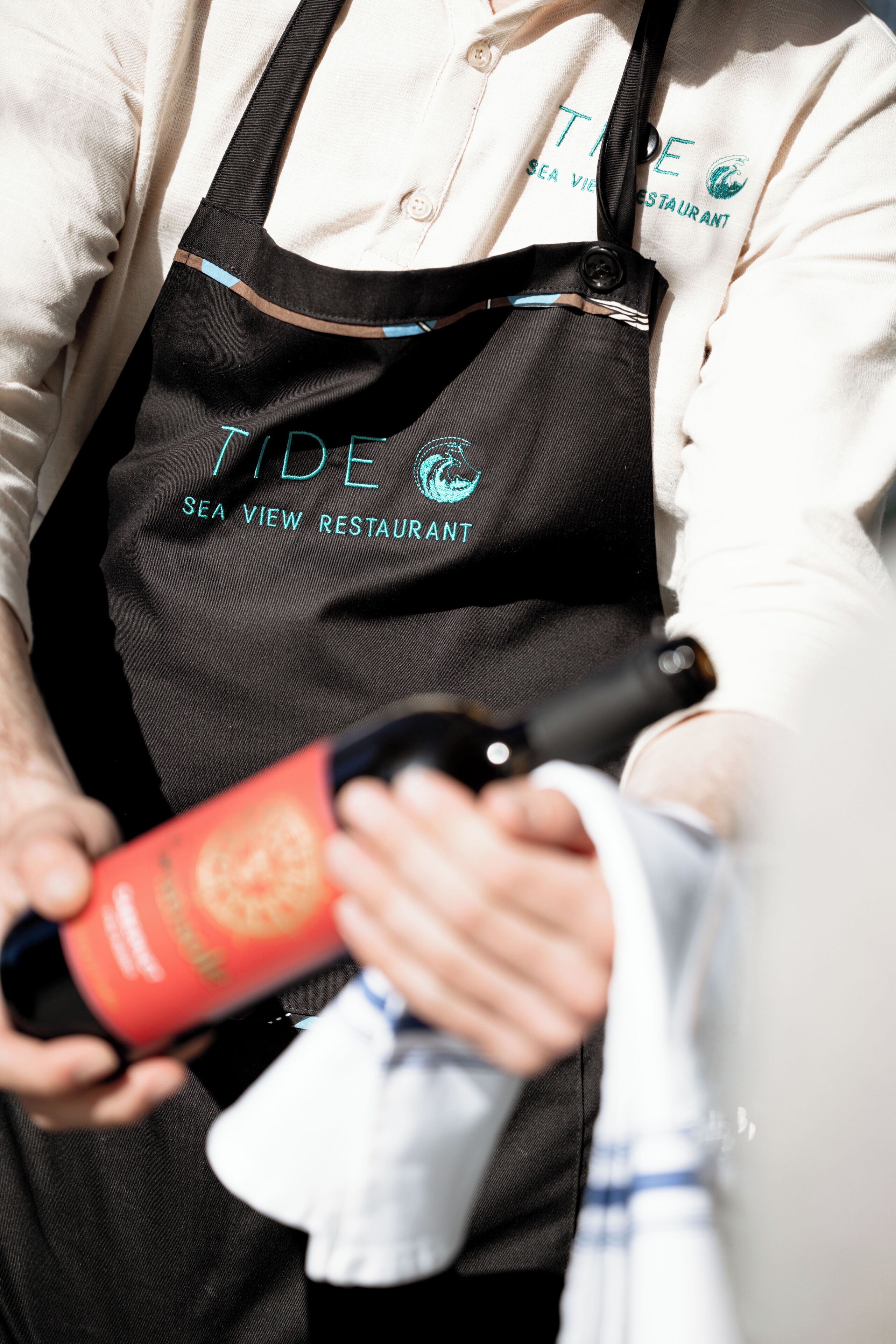 A person wearing a black apron with "TIDE SEA VIEW RESTAURANT" printed on it, holding a bottle.
