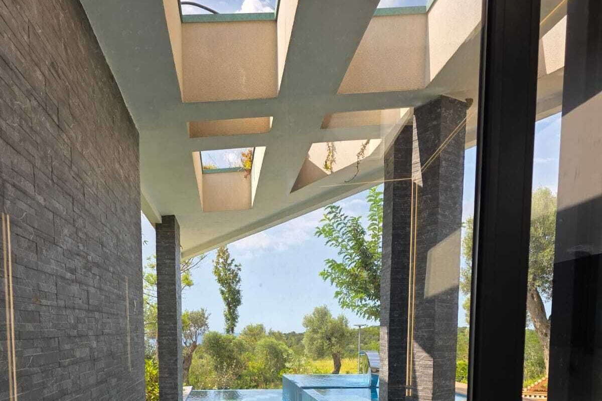 A modern indoor swimming pool with clear water, surrounded by stone walls and glass doors, offering views of greenery and a clear sky.