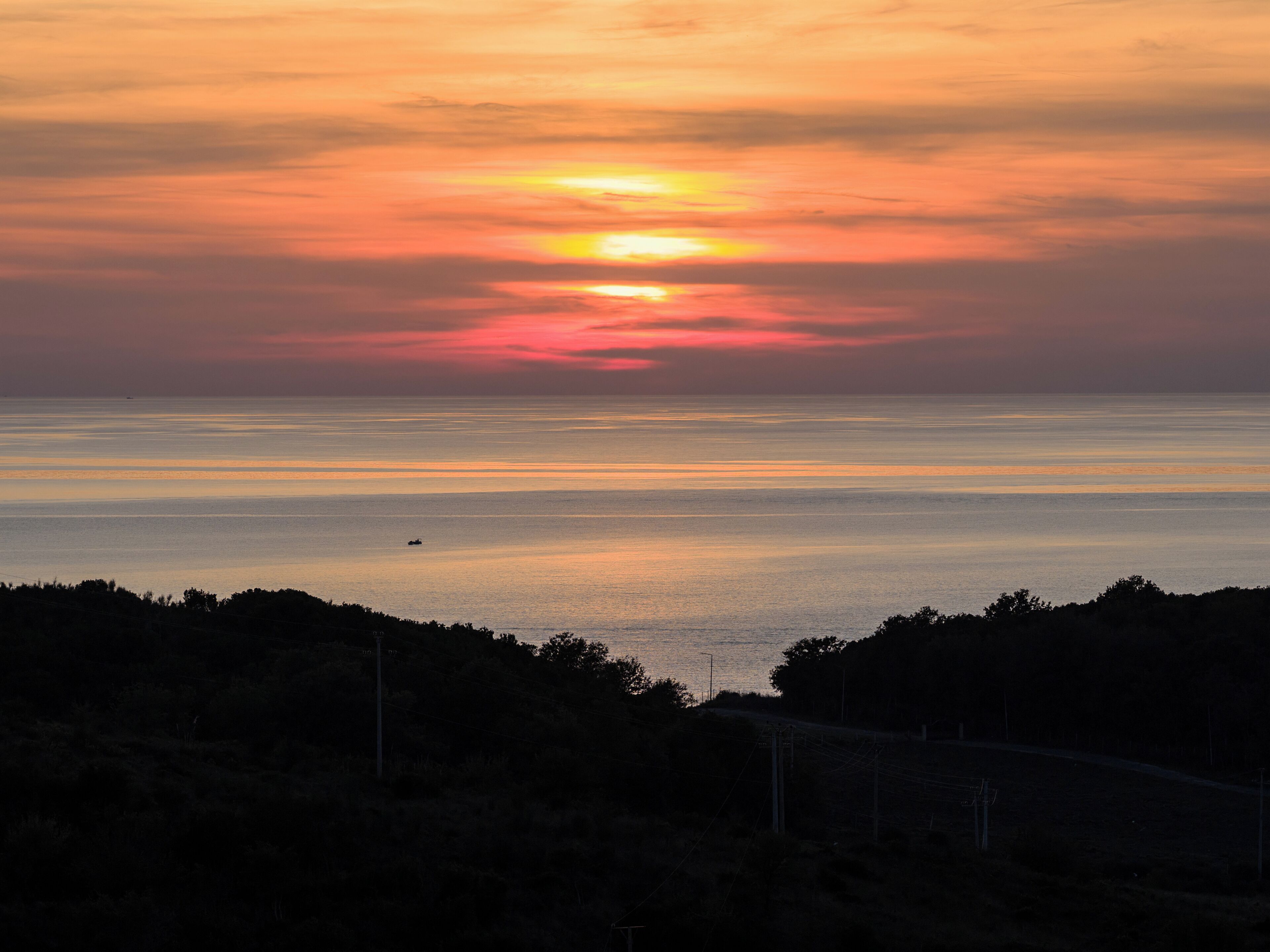 A sunset over a calm sea with a silhouette of trees and a distant boat.