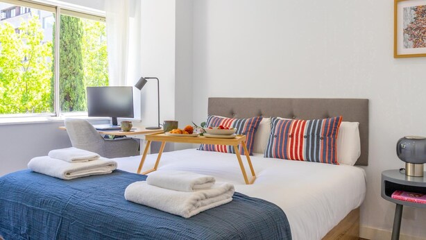 Ein modernes Hotelzimmer mit einem großen Bett, einem Schreibtisch mit Laptop, einem Nachttisch mit Lampe und Blick ins Grüne.