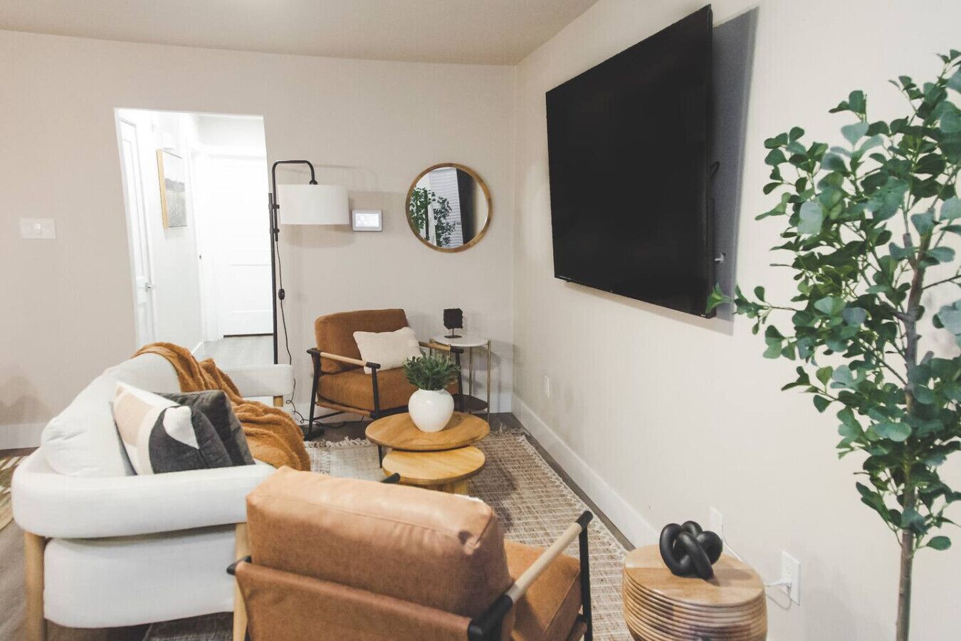 A living room with a white sofa, a brown armchair, a coffee table, a TV mounted on the wall, a mirror, and a potted plant.