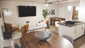 A modern living room with a wooden dining table, a sofa, and a kitchen area with a foosball table. - Fort Worth 2BR/2BA Home w/ Pool + Games | Sleeps 6 (Fort Worth)
