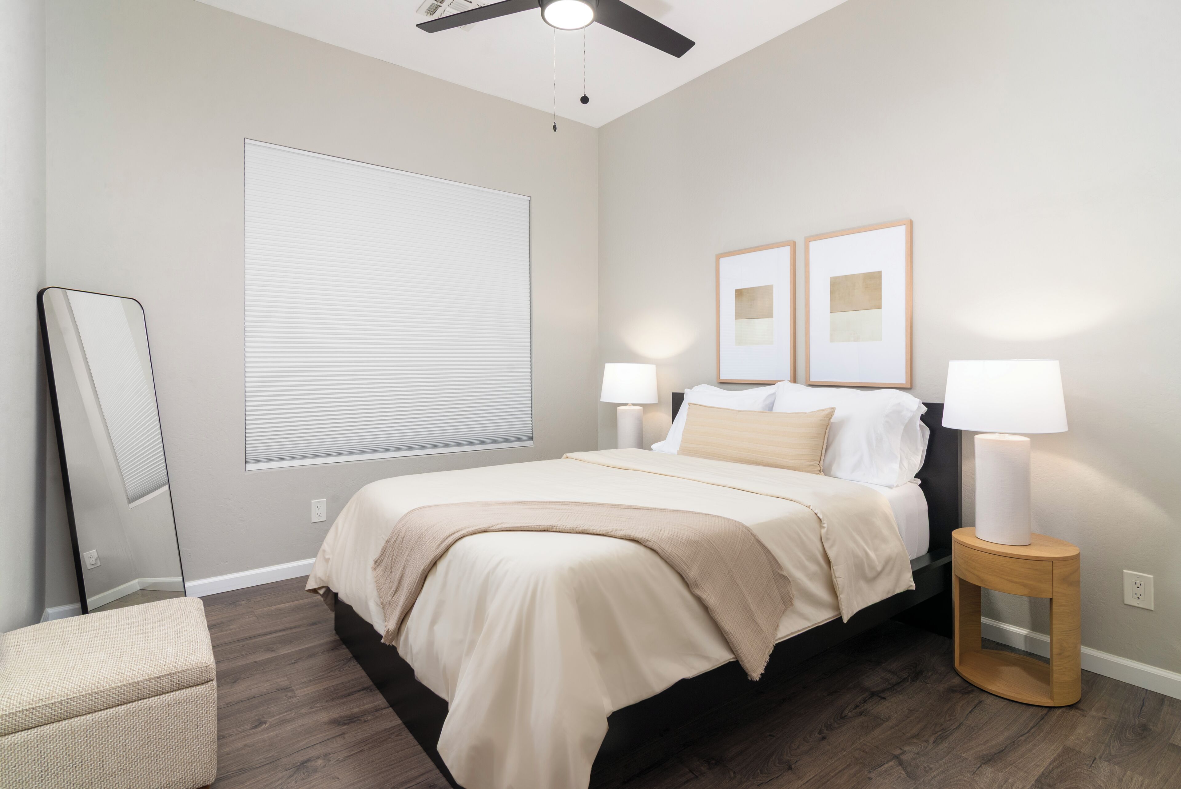 Une chambre à coucher comprenant un lit, des lampes de chevet, un ventilateur de plafond, une grande fenêtre avec des stores et deux tableaux encadrés accrochés au mur.