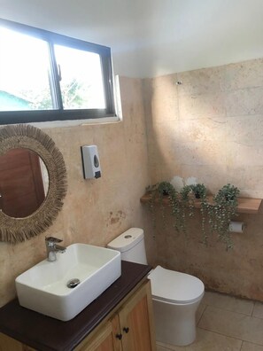 A bathroom with a rectangular sink, a toilet, a mirror with a woven frame, and a window above the sink.