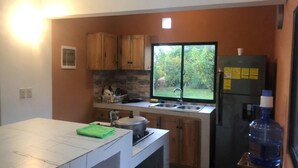 A compact kitchen with wooden cabinets, a built-in stove, a sink, and a window with a view of greenery. - Casa Playa Rincon Hills Samana (Samaná)