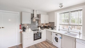 A modern kitchen with white cabinetry, a built-in oven, microwave, and washing machine, a stainless steel range hood, and a window with blinds. - Spacious country suite located at Lonan Country House & Gardens in Scotland! (Taynuilt)