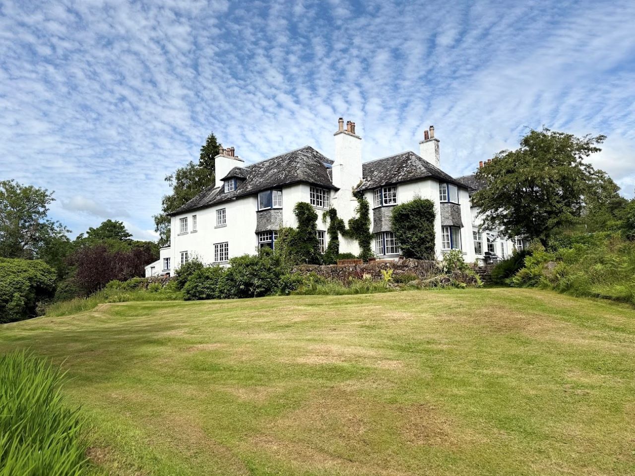 A large white building with multiple chimneys and windows, surrounded by a grassy lawn and trees.
