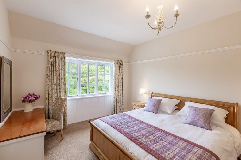A bedroom with a wooden bed, a bedside table, a chair, a window with curtains, and a chandelier.